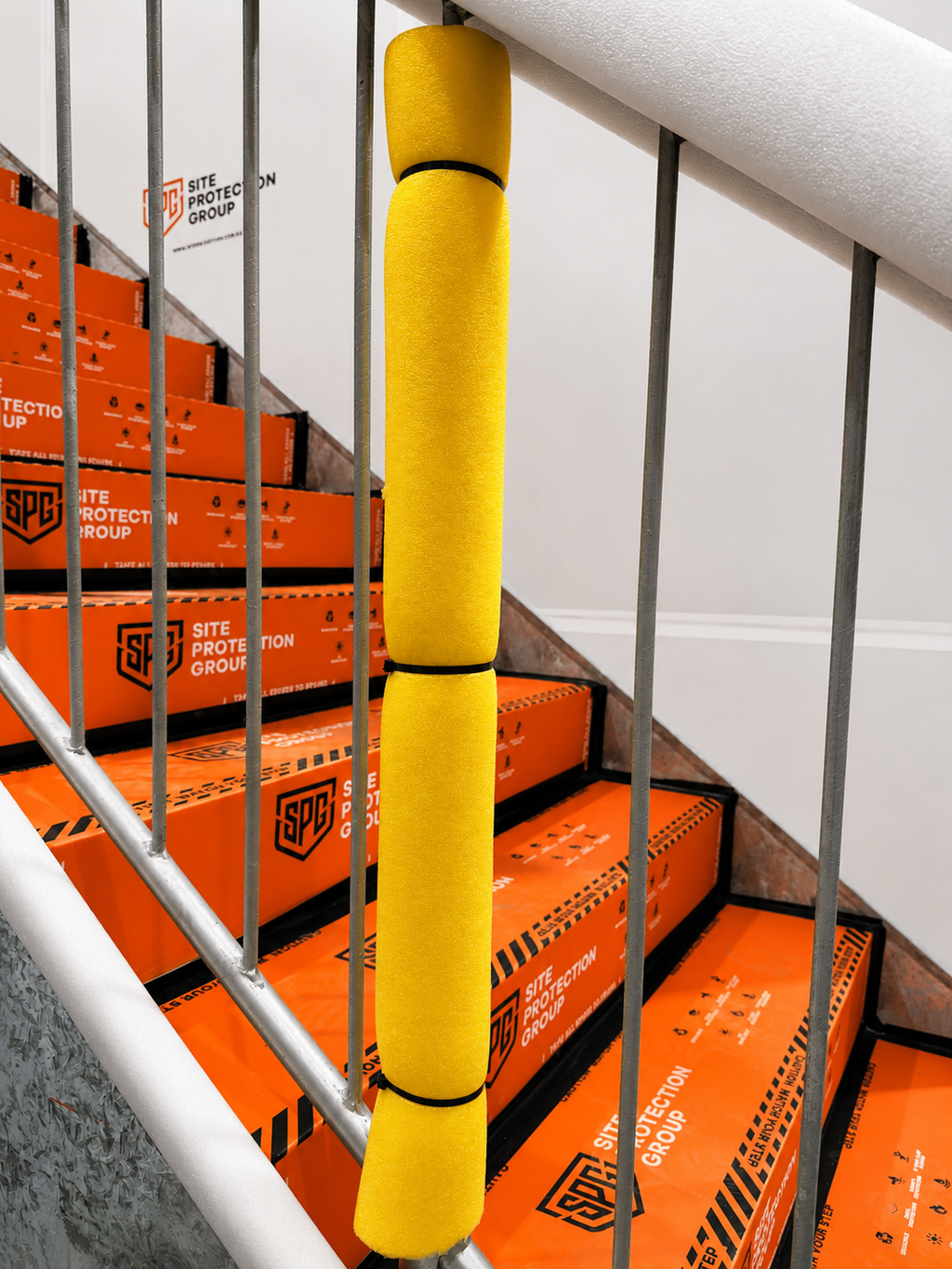 A bright yellow Site Protection Group anti-slip stair tape wrapped around a staircase railing, with orange branded safety boxes stacked on the steps behind it.