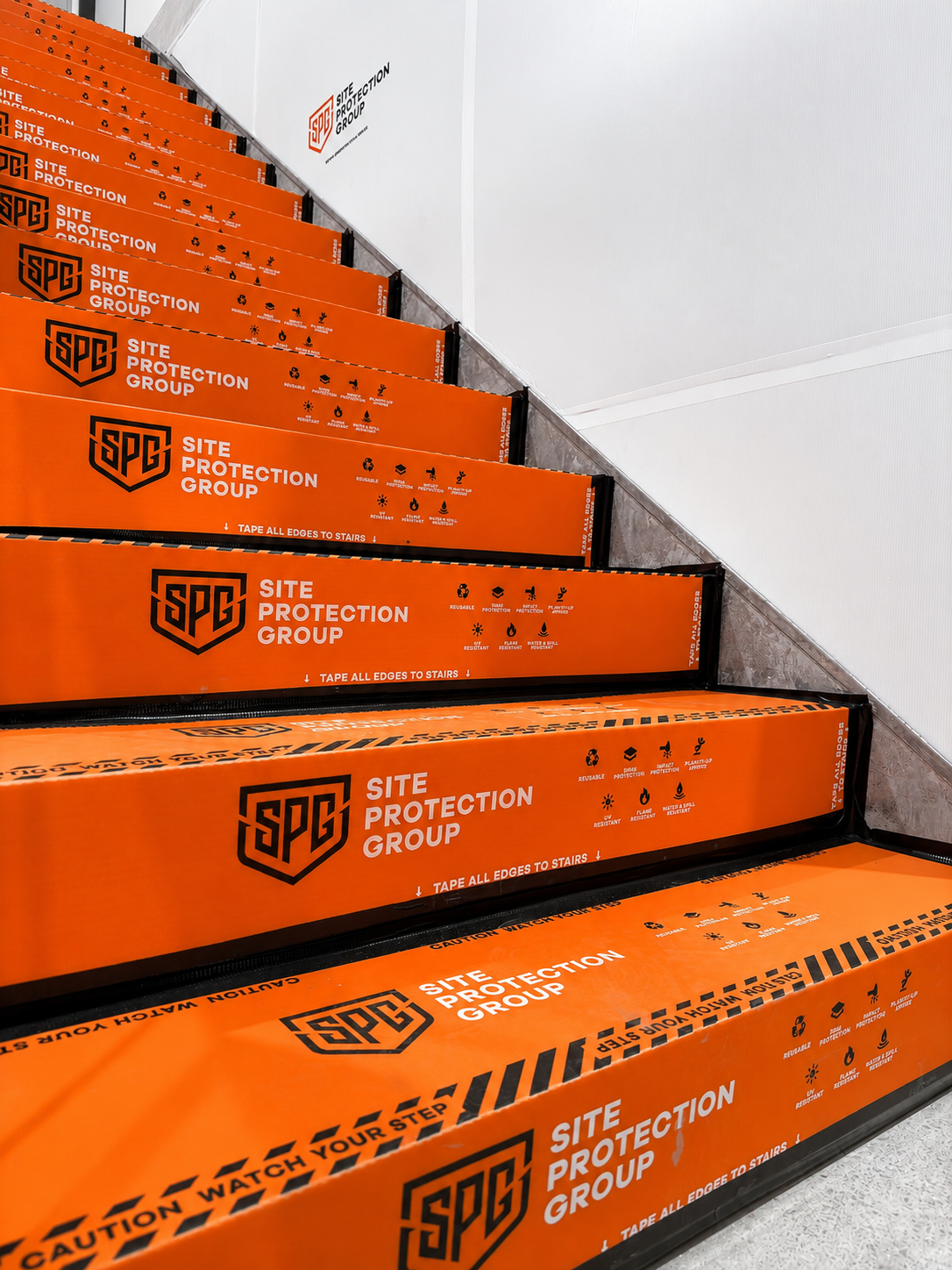 Bright orange stair treads wrapped with repeated Site Protection Group branding and caution-style graphics, installed on an indoor staircase against a white wall.
